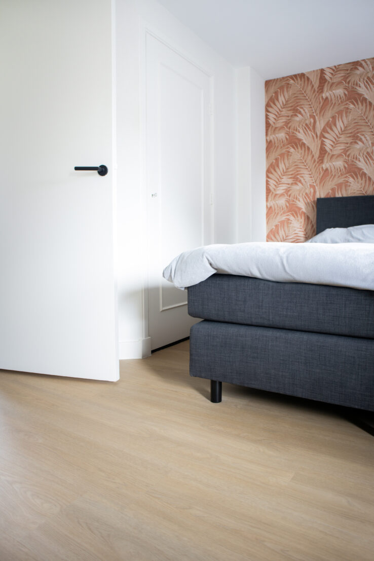 Moderne slaapkamer met een lichte PVC-vloer, een grijs bed en een beige muur met een tropisch patroon.