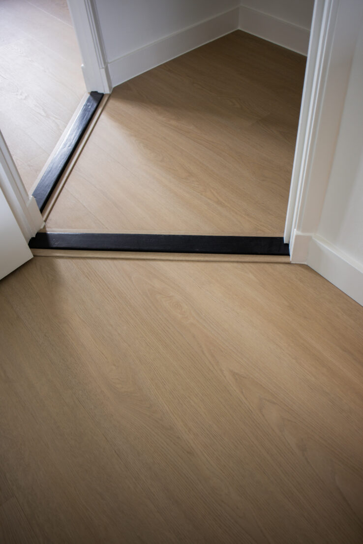 Lichte houtlook PVC-vloer bij twee deuropeningen, met zwarte overgangsstrips die de ruimtes met elkaar verbinden.