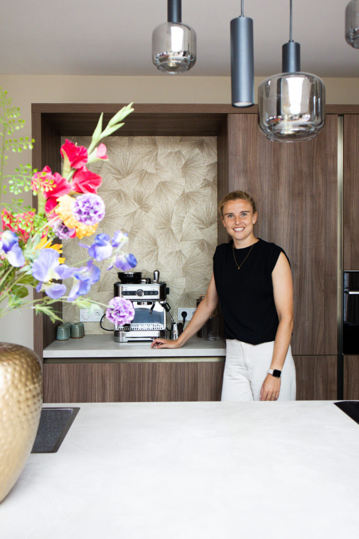 Vrouw in een moderne keuken die naast een espressomachine staat, met houten kasten, lichte werkbladen en kleurrijke bloemen op de voorgrond.