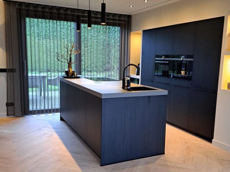 Lichte eiken PVC-vloer in een moderne keuken met een centraal kookeiland, zwarte keukenkastjes en grote ramen voor veel natuurlijk licht.