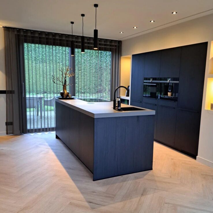 Lichte eiken PVC-vloer in een moderne keuken met een centraal kookeiland, zwarte keukenkastjes en grote ramen voor veel natuurlijk licht.