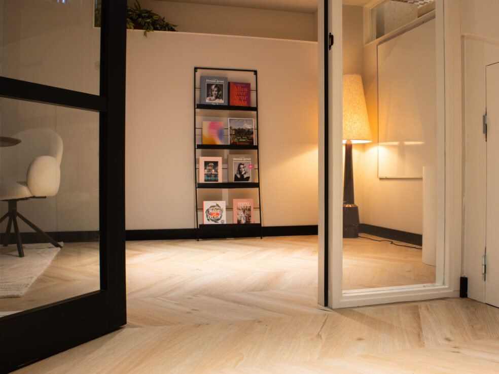 Lichte eiken PVC-vloer in een moderne ruimte met een glazen wand, een boekenplank en een stijlvolle bureaustoel.