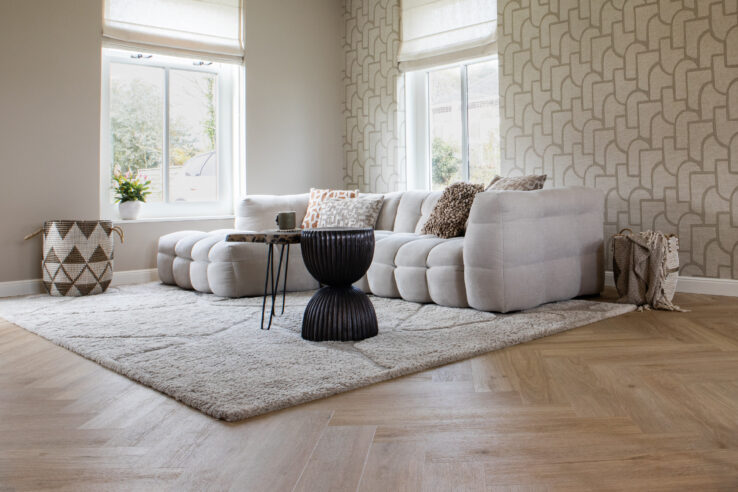 Lichte visgraat PVC-vloer in een warme woonkamer met een zachte beige hoekbank, hoogpolig vloerkleed en grafisch behang.