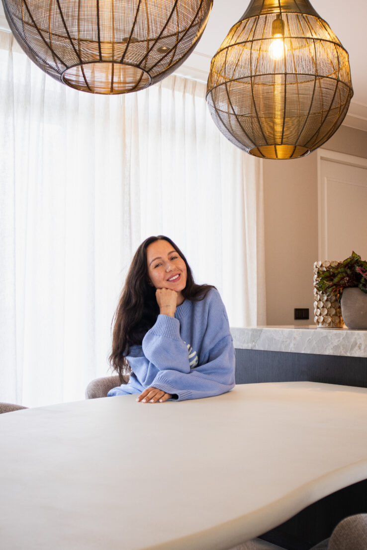 Vrouw die lachend aan een lichte eettafel zit, met twee grote rotan hanglampen boven haar en zachte, lichte gordijnen op de achtergrond.