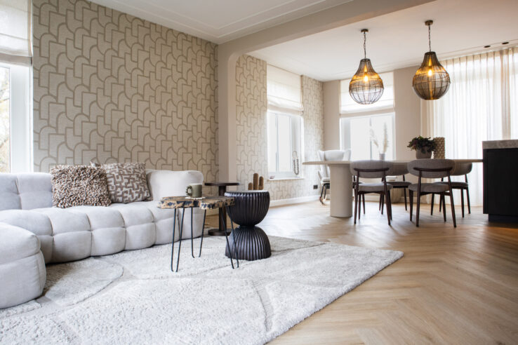 Moderne woonkamer met een lichtgekleurde bank, een vloerkleed, en een houten visgraatvloer. De ruimte heeft grote ramen met veel natuurlijk licht, stijlvolle verlichtingsarmaturen en een eethoek met houten stoelen.