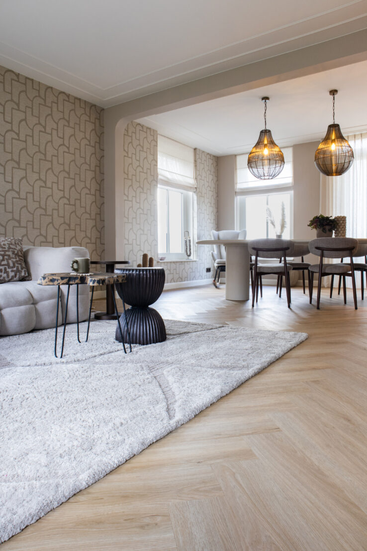 Woonkamer met lichte eiken walvisgraatvloer, een groot zacht vloerkleed, een beige bank en een eettafel met moderne donkere stoelen, afgewerkt met twee grote hanglampen en grafisch behang.