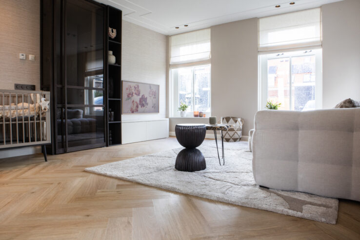 Lichte visgraat PVC-vloer in een moderne woonkamer met hoogpolig vloerkleed, beige bank, donkere kastwand en grote ramen met veel daglicht.