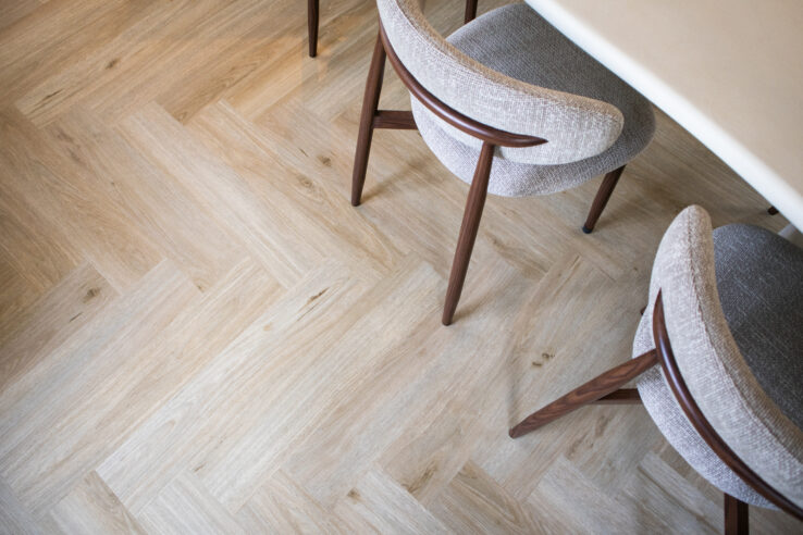 Detail van een lichte visgraat PVC-vloer met eetkamerstoelen van hout en stof rond een lichte tafel.