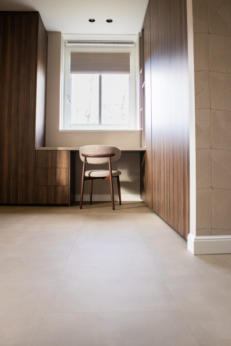 Minimalistische werkruimte met een houten bureau, een stoel en een raam met uitzicht op de natuur. De vloer is bedekt met grote, neutrale tegels en de wanden zijn afgewerkt met houten panelen.