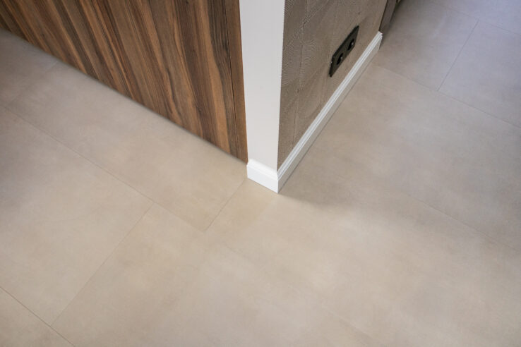 Detail van een lichte, beige tegel-look PVC-vloer naast een houtlook kast en witte plint.