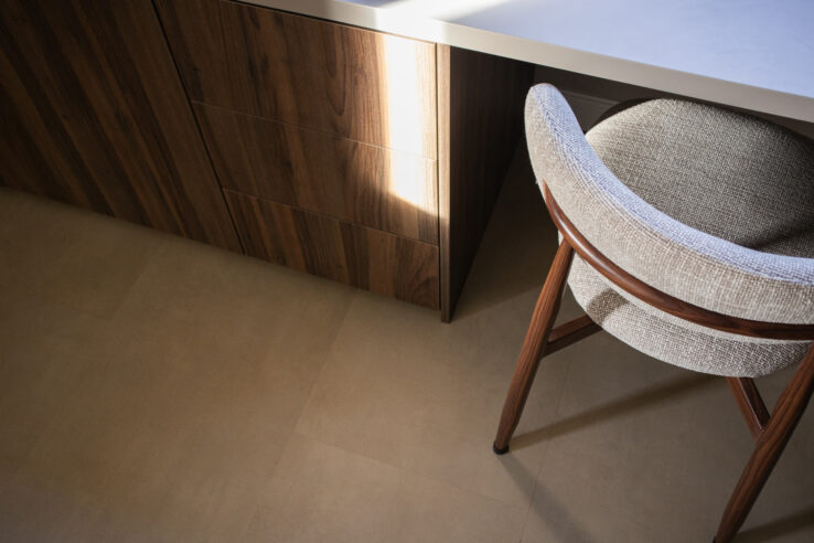 Detail van een beige tegel-look PVC-vloer naast een donkere houtlook kast en een gestoffeerde stoel met houten poten, belicht door invallend zonlicht.