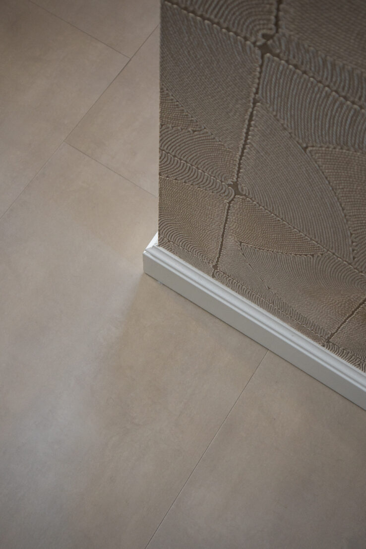 Detailfoto van een beige PVC-tegelvloer naast een wand met reliëfstructuur en een witte plint.
